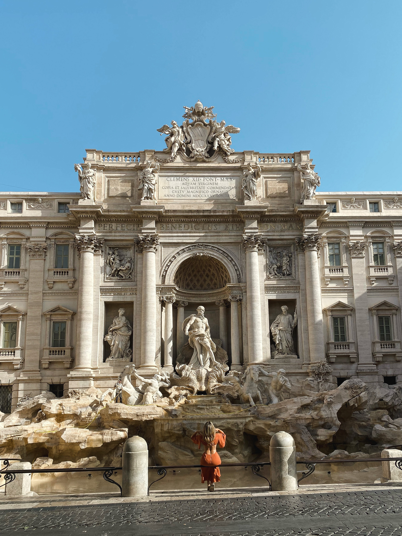 A visitor in an orange dress stands in awe before the imposing Trevi Fountain, an iconic baroque masterpiece in the heart of Rome, basking in the glow of the sun as seen in slide7