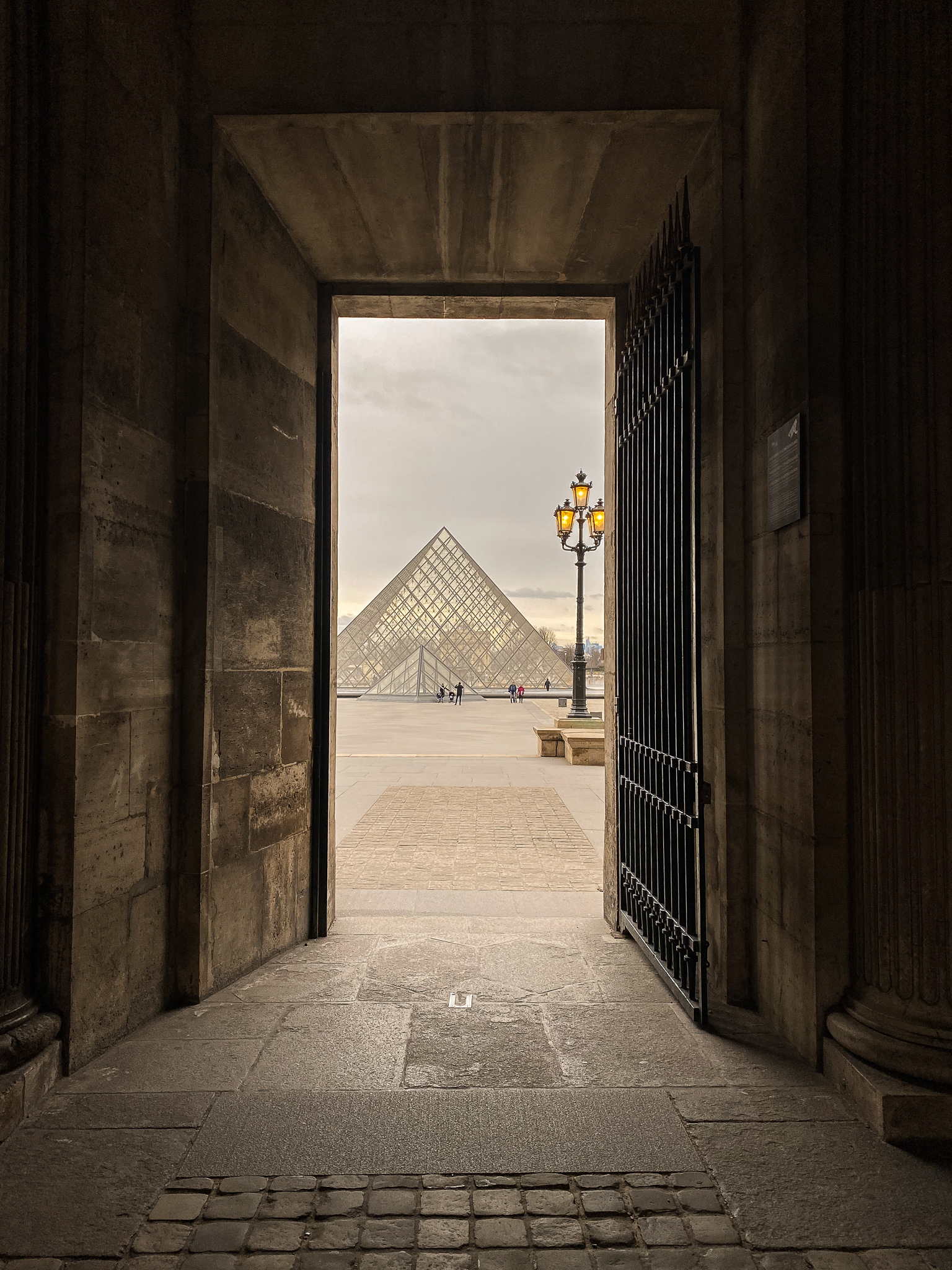 A creative header: A view from a shadowed archway frames the iconic Louvre Pyramid in the distance, with the soft glow of a streetlamp and the warm tones of the surrounding architecture inviting observers